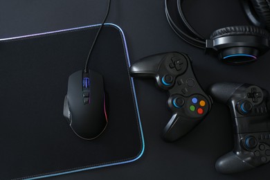 Computer mouse, mousepad, headset and game controllers on black background, flat lay