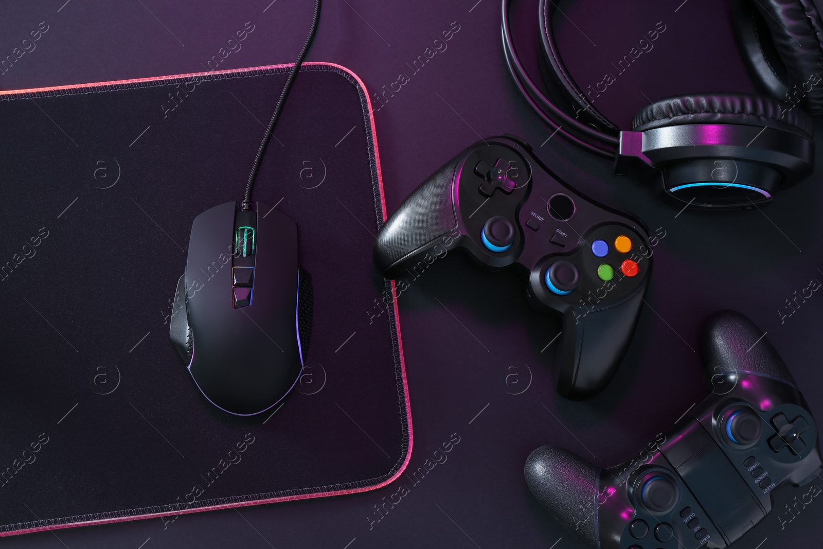 Photo of Computer mouse, mousepad, headset and game controllers in neon lights on black background, flat lay