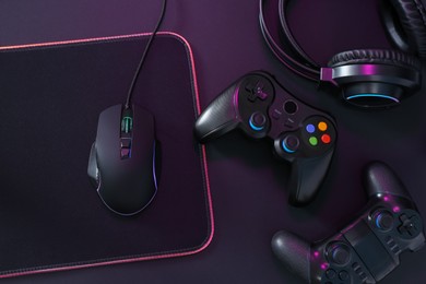 Photo of Computer mouse, mousepad, headset and game controllers in neon lights on black background, flat lay