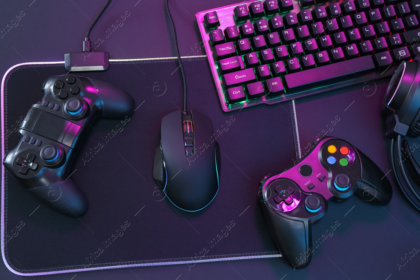 Photo of Computer mouse, RGB keyboard and game controllers in neon lights on dark background, top view