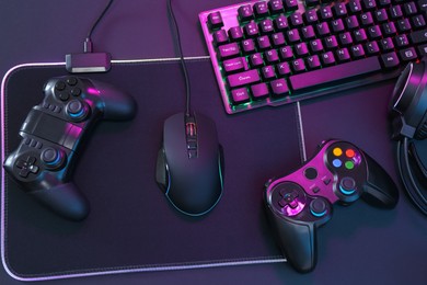 Computer mouse, RGB keyboard and game controllers in neon lights on dark background, top view