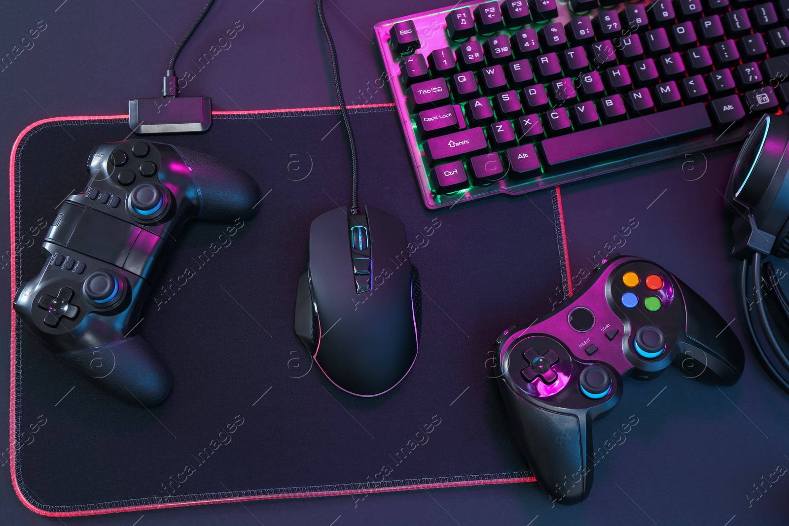 Photo of Computer mouse, RGB keyboard and game controllers in neon lights on dark background, top view