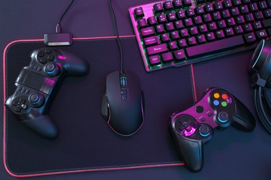 Computer mouse, RGB keyboard and game controllers in neon lights on dark background, top view