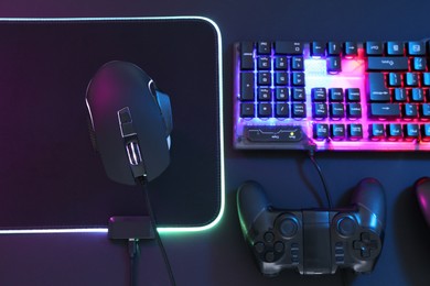 Photo of Computer mouse, RGB keyboard and game controller in neon lights on dark background, flat lay