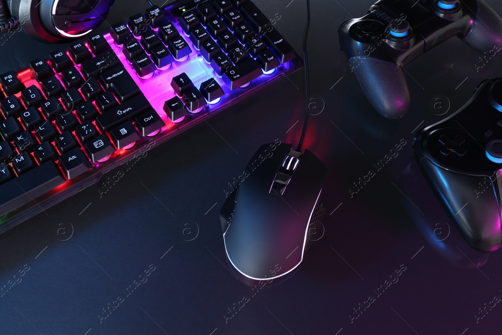 Photo of Computer mouse, RGB keyboard and game controllers in neon lights on dark background, above view