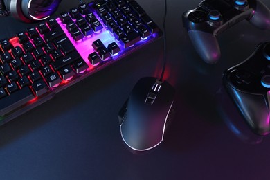 Computer mouse, RGB keyboard and game controllers in neon lights on dark background, above view