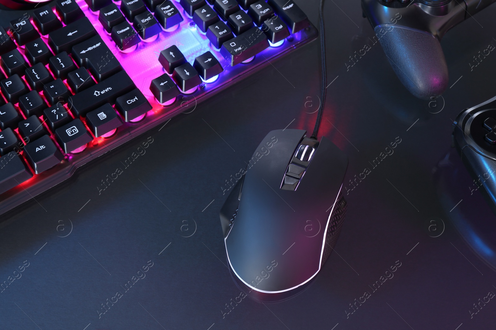 Photo of Computer mouse, RGB keyboard and game controllers in neon lights on dark background, above view