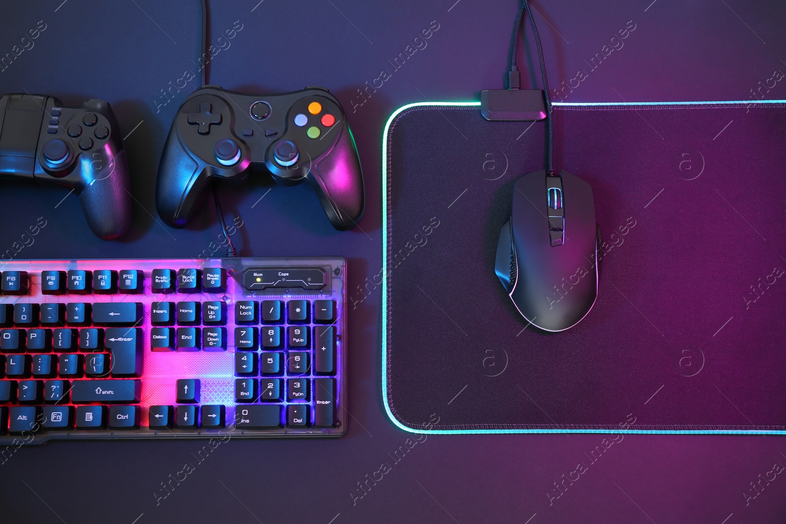 Photo of Computer mouse, RGB keyboard and game controllers in neon lights on dark background, flat lay