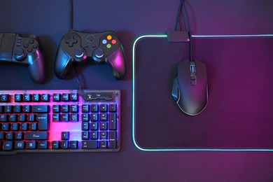 Computer mouse, RGB keyboard and game controllers in neon lights on dark background, flat lay