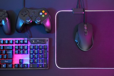 Photo of Computer mouse, RGB keyboard and game controllers in neon lights on dark background, flat lay