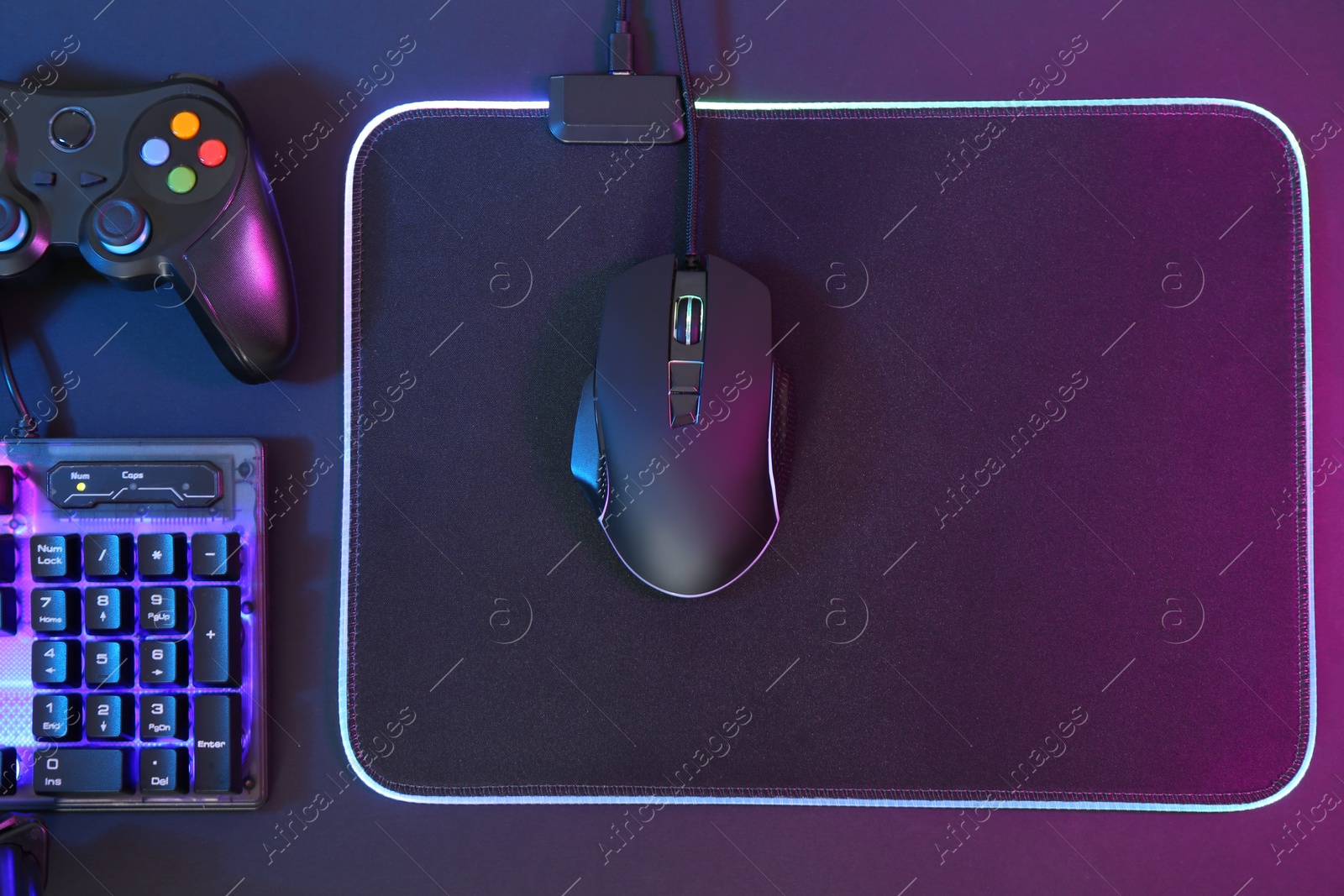 Photo of Computer mouse, RGB keyboard and game controller in neon lights on dark background, flat lay