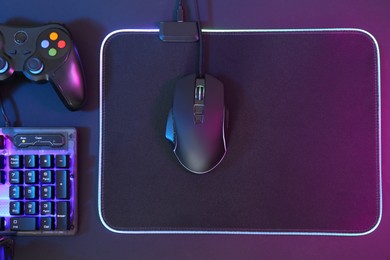 Photo of Computer mouse, RGB keyboard and game controller in neon lights on dark background, flat lay