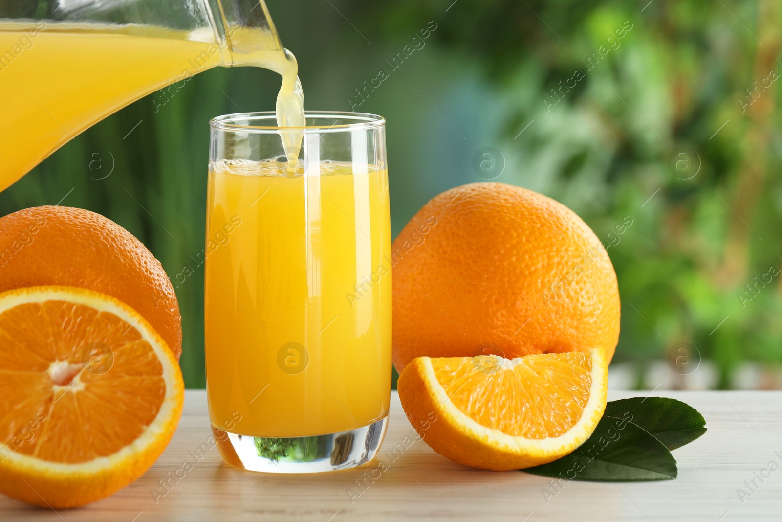 Photo of Pouring tasty orange juice into glass at white wooden table outdoors