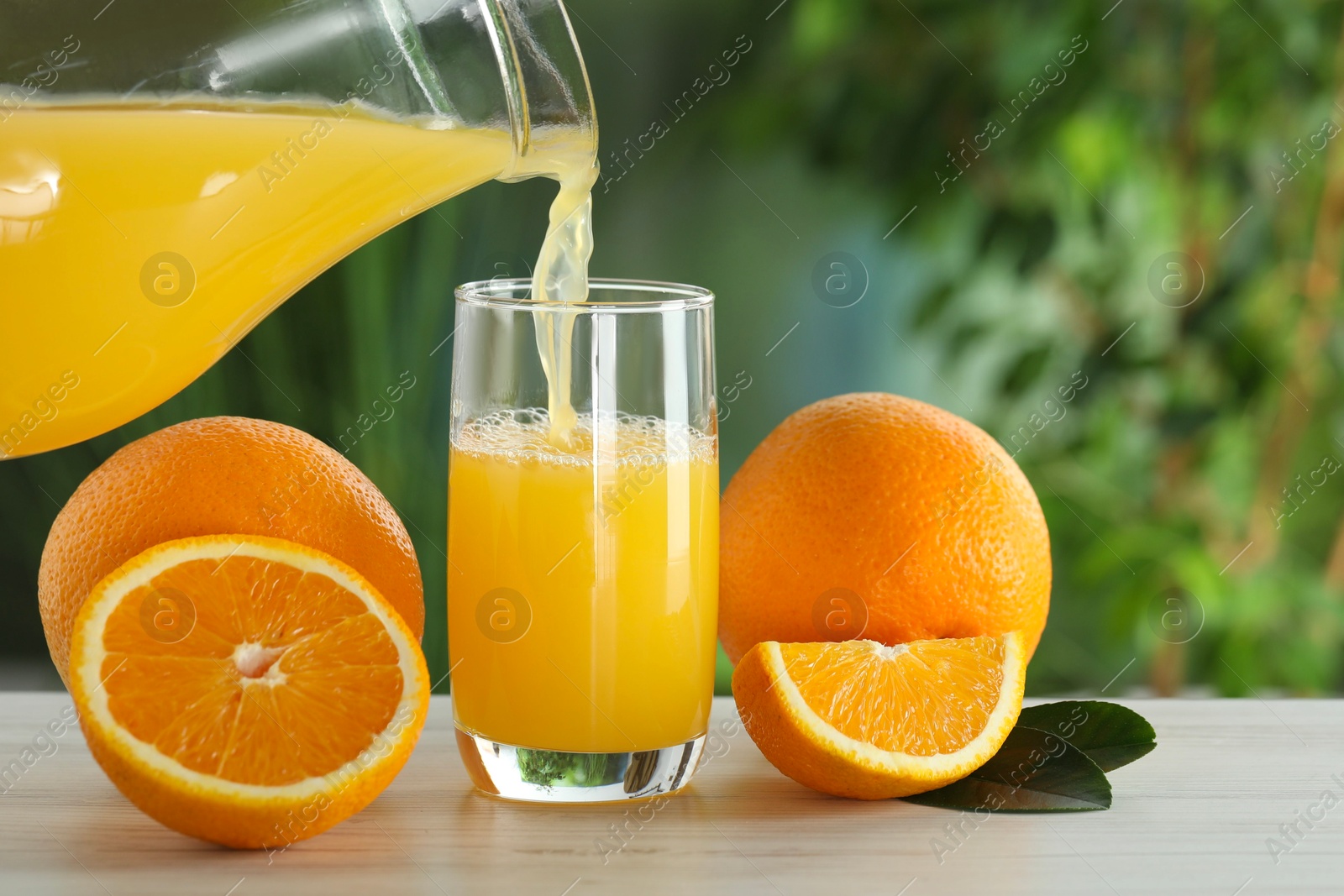 Photo of Pouring tasty orange juice into glass at white wooden table outdoors