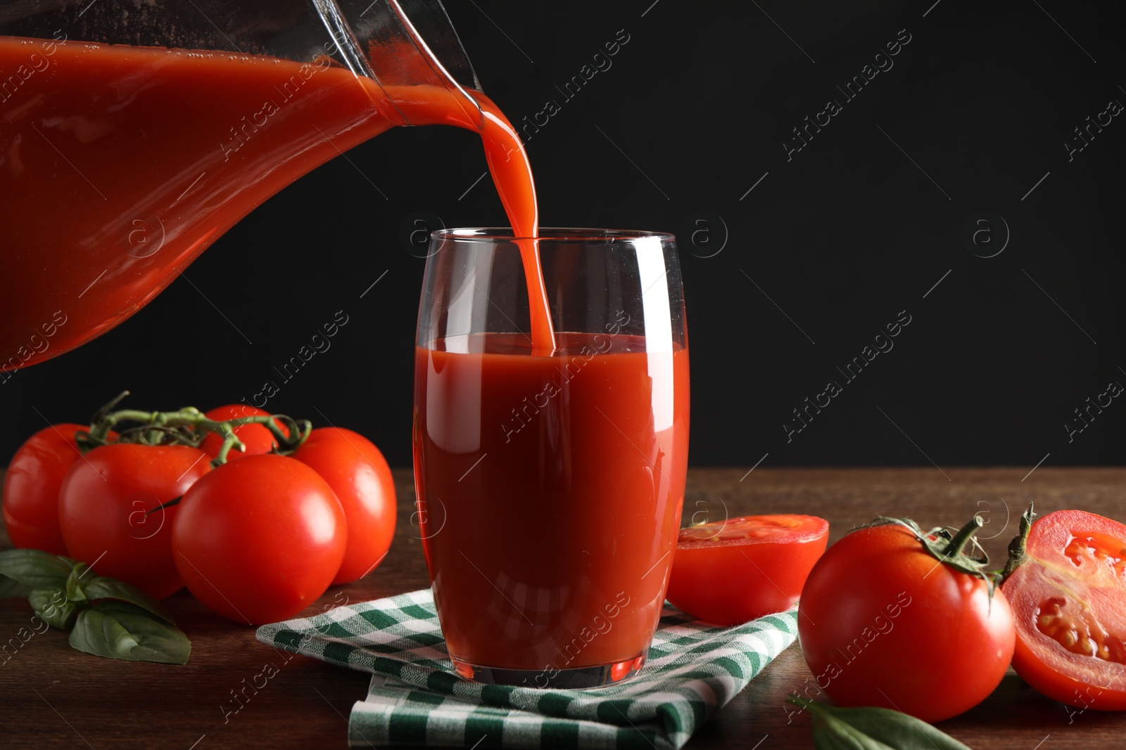 Photo of Pouring tasty tomato juice from jug into glass at wooden table with vegetables