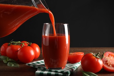 Pouring tasty tomato juice from jug into glass at wooden table with vegetables