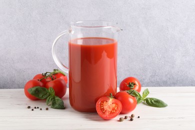 Photo of Tasty tomato juice in jug, basil leaves, peppercorns and vegetables on white wooden table indoors