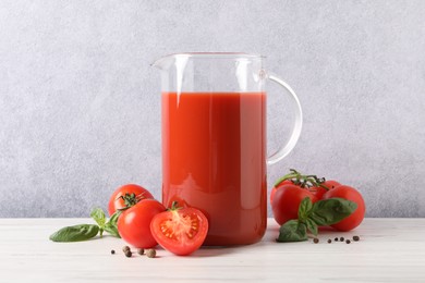 Photo of Tasty tomato juice in jug, basil leaves, peppercorns and vegetables on white wooden table indoors