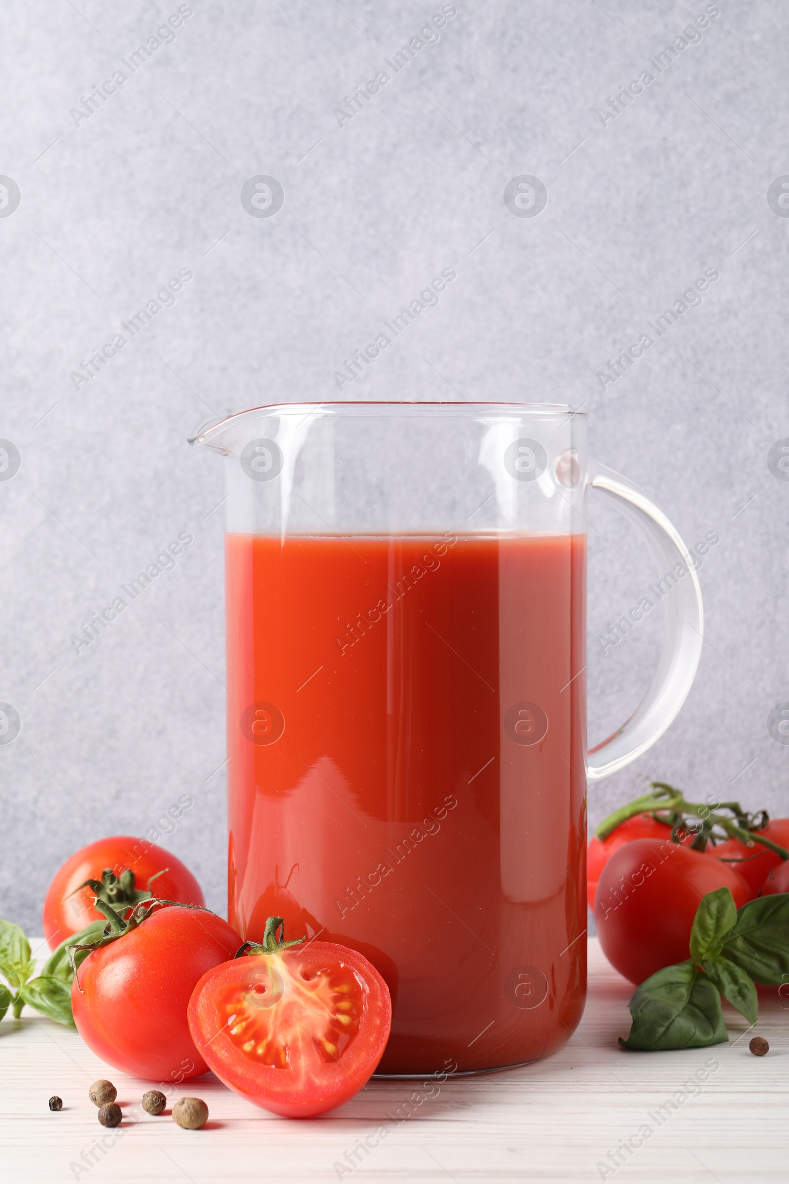 Photo of Tasty tomato juice in jug, basil leaves, peppercorns and vegetables on white wooden table indoors