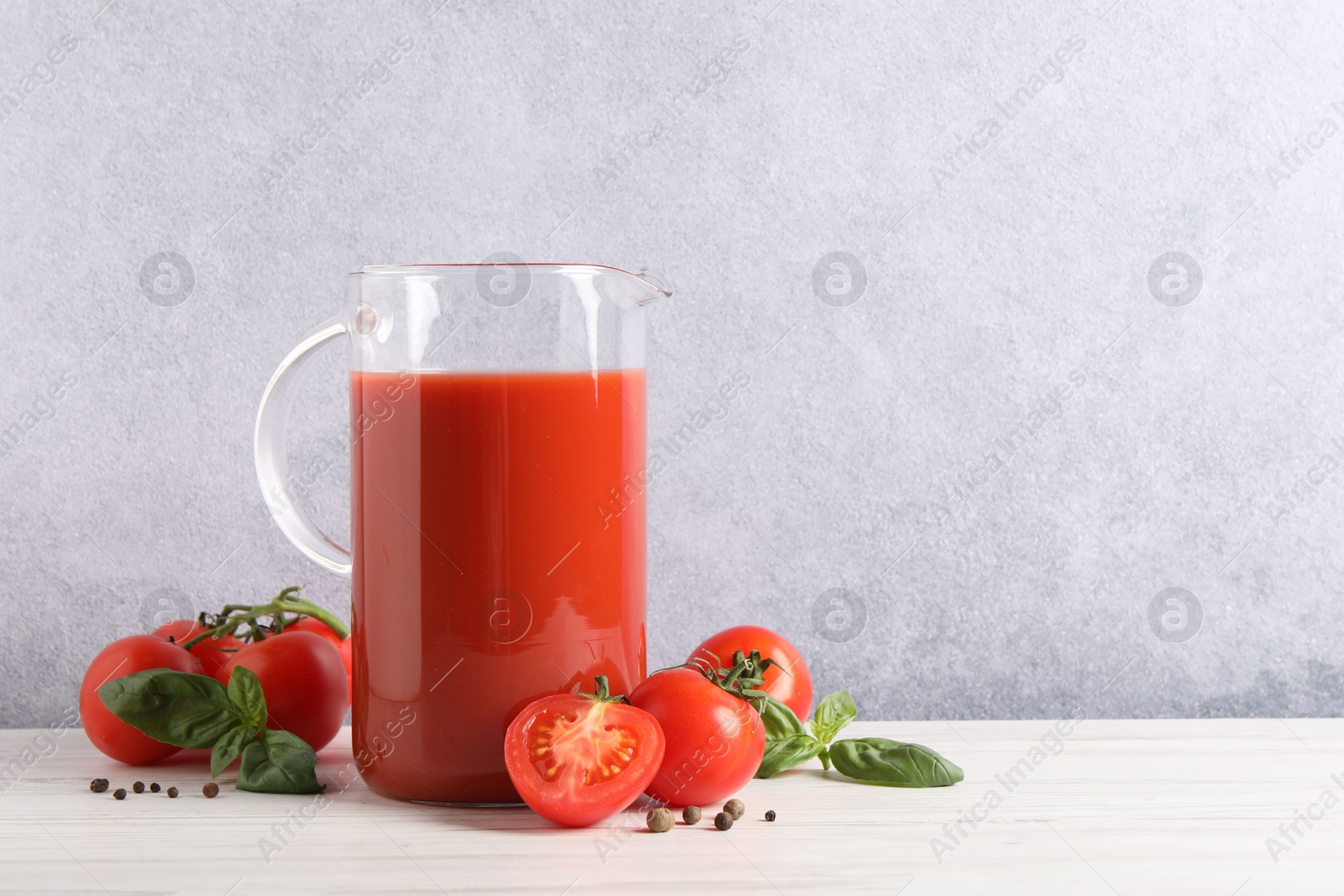 Photo of Tasty tomato juice in jug, basil, peppercorns and vegetables on white wooden table indoors. Space for text