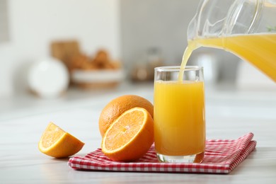 Pouring tasty orange juice from jug into glass at white table with citrus fruits