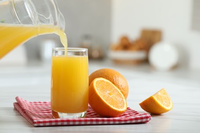 Pouring tasty orange juice from jug into glass at white table with citrus fruits