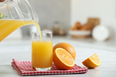 Pouring tasty orange juice from jug into glass at white table with citrus fruits