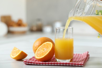 Pouring tasty orange juice from jug into glass at white table with citrus fruits