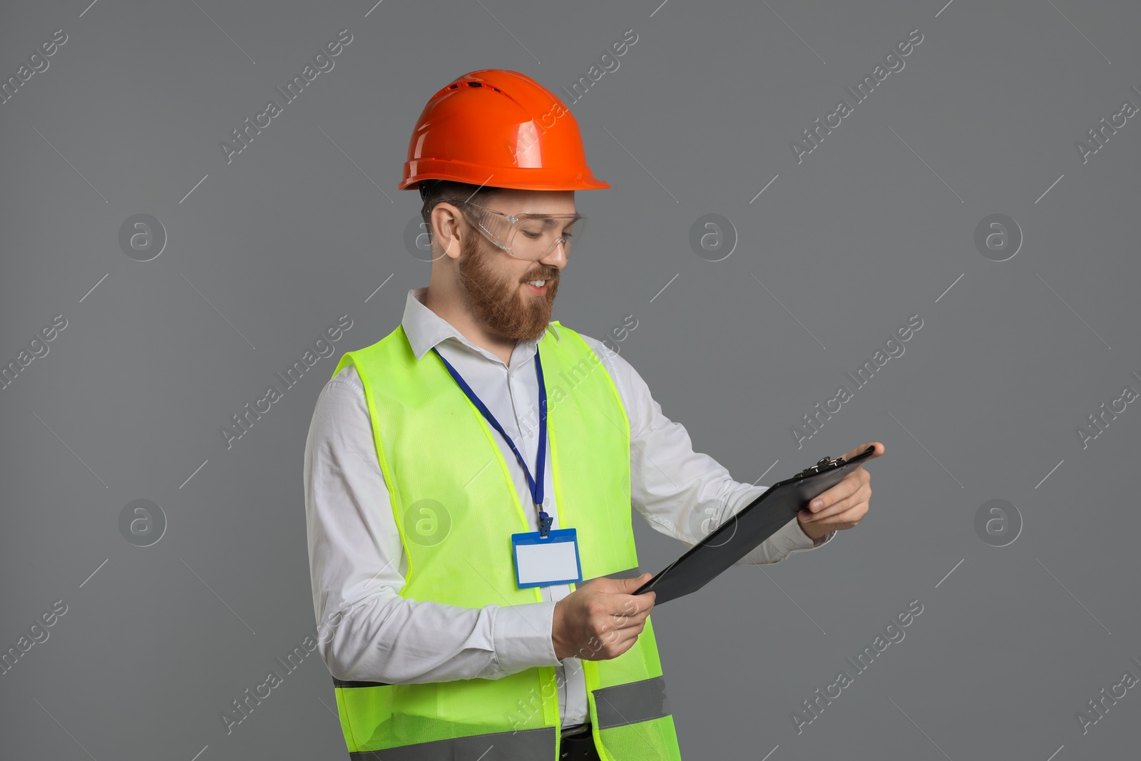 Photo of Engineer in hard hat and goggles holding clipboard on grey background