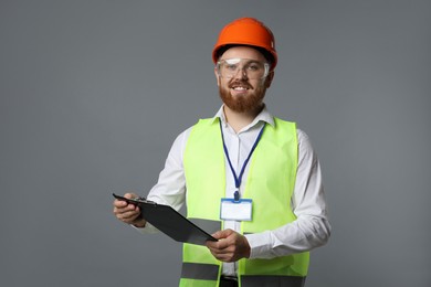 Photo of Engineer in hard hat and goggles holding clipboard on grey background