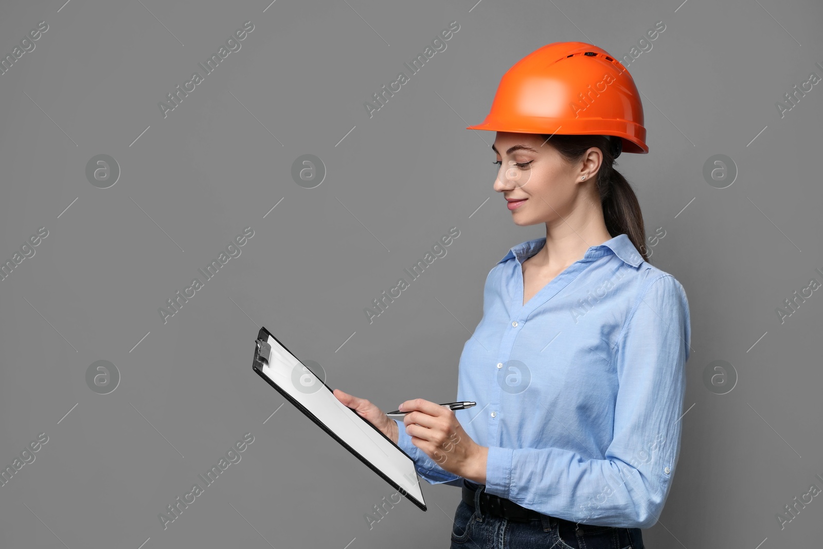 Photo of Engineer in hard hat with clipboard and pen on grey background, space for text