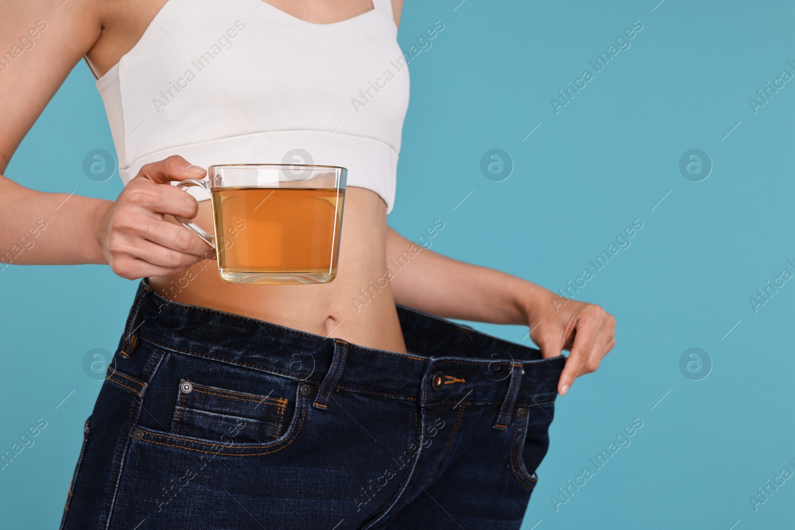 Photo of Weight loss concept. Woman with cup of diet tea and big pants on light blue background, closeup