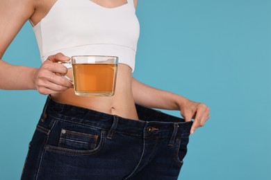 Photo of Weight loss concept. Woman with cup of diet tea and big pants on light blue background, closeup