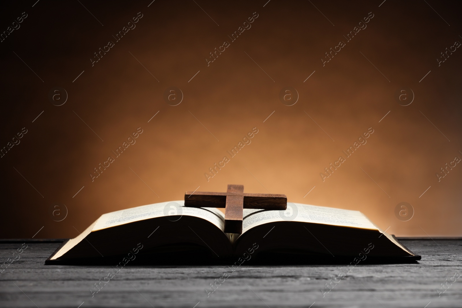 Photo of Bible and cross on gray wooden table against brown background, space for text. Religion of Christianity