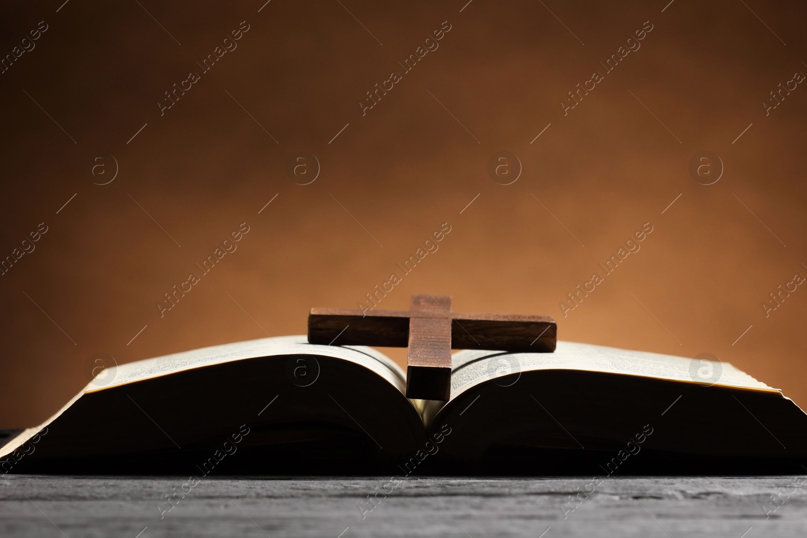 Photo of Bible and cross on gray table against brown background, space for text. Religion of Christianity