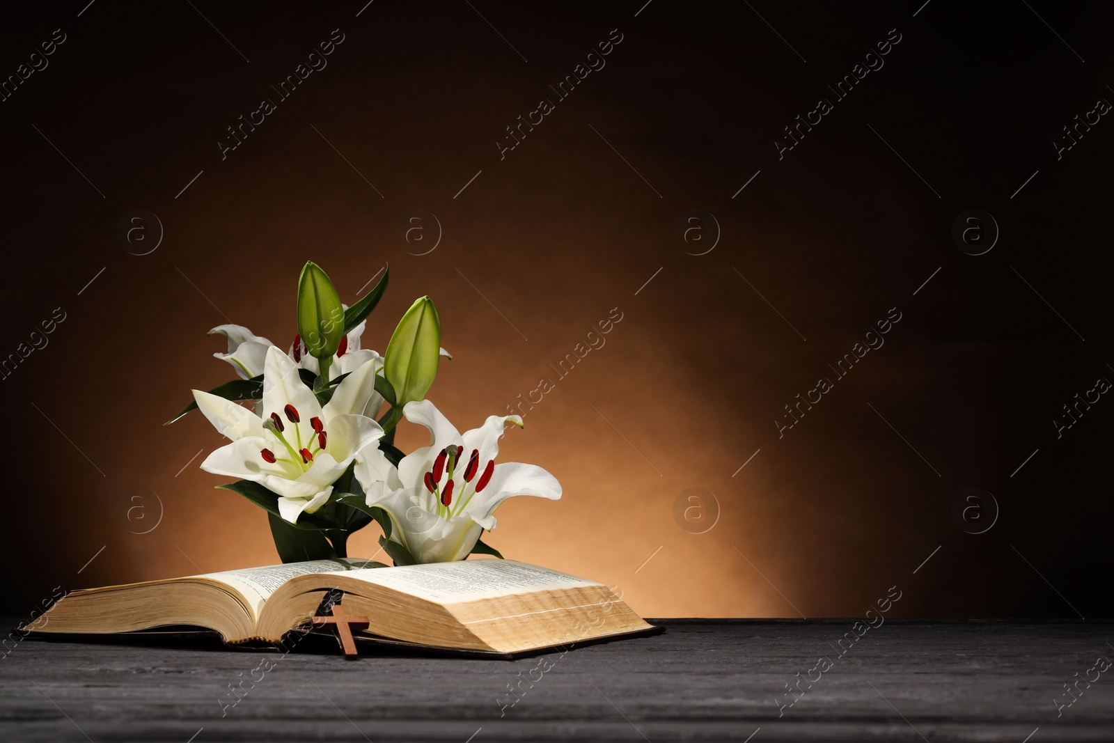 Photo of Bible, cross and lilies on gray table against brown background, space for text. Religion of Christianity