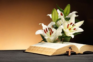 Photo of Bible, cross and lilies on gray table against brown background, space for text. Religion of Christianity