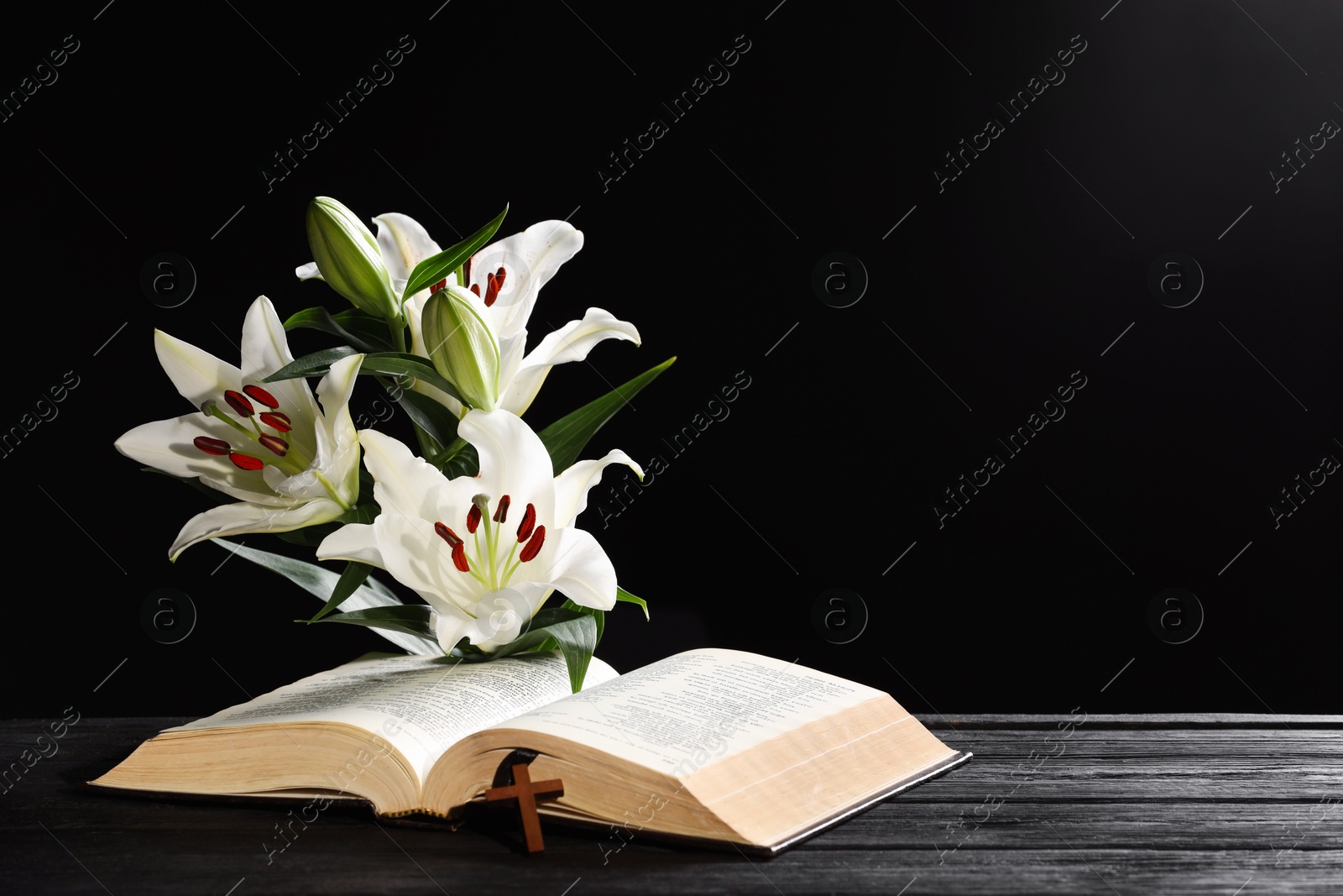 Photo of Bible, cross and lilies on dark gray wooden table against black background, space for text. Religion of Christianity