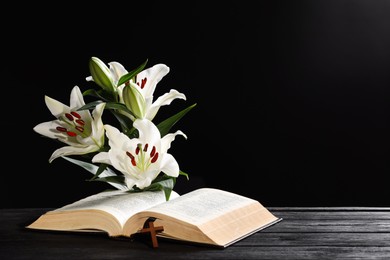 Bible, cross and lilies on dark gray wooden table against black background, space for text. Religion of Christianity