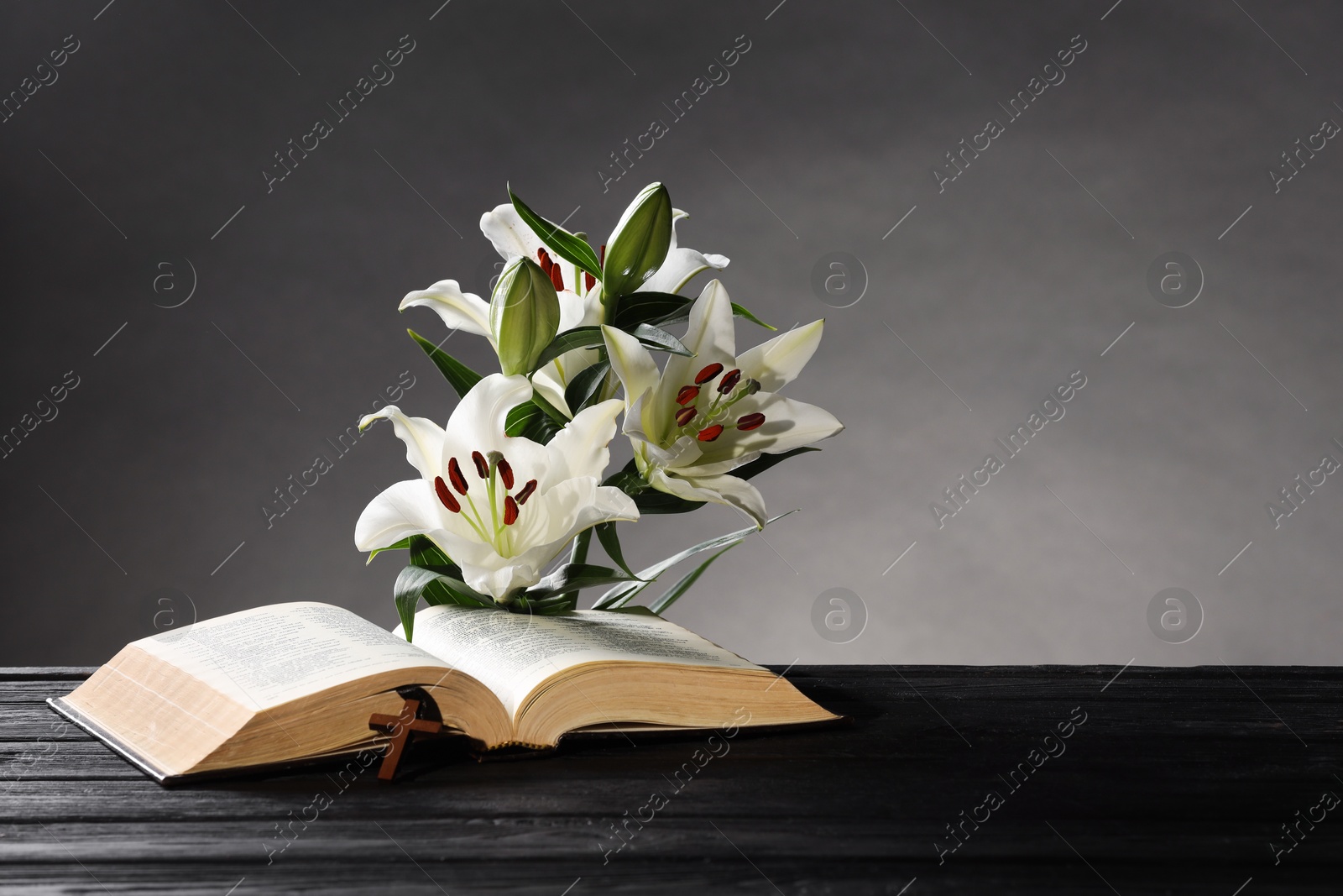 Photo of Bible, cross and lilies on dark wooden table against gray background, space for text. Religion of Christianity