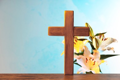 Photo of Wooden cross and beautiful lily flowers on table against light blue background, space for text. Religion of Christianity