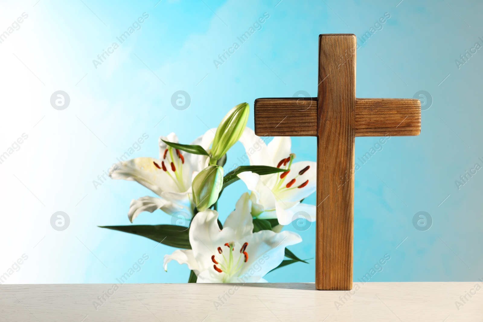 Photo of Wooden cross and beautiful lily flowers on table against light blue background, space for text. Religion of Christianity