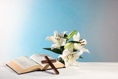 Photo of Wooden cross, Bible and beautiful lily flowers on white table against light blue background, space for text. Religion of Christianity