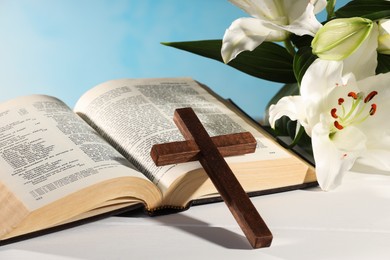 Wooden cross, Bible and beautiful lily flowers on white table against light blue background, closeup. Religion of Christianity