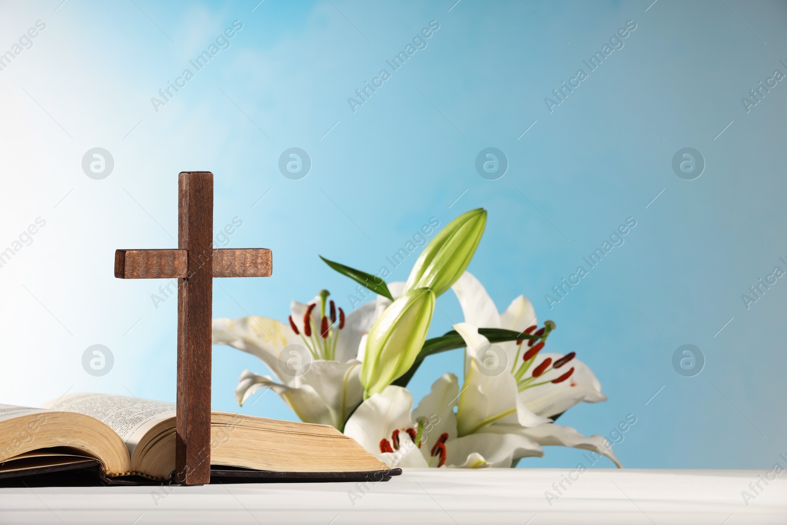 Photo of Wooden cross, Bible and beautiful lily flowers on white table against light blue background, space for text. Religion of Christianity