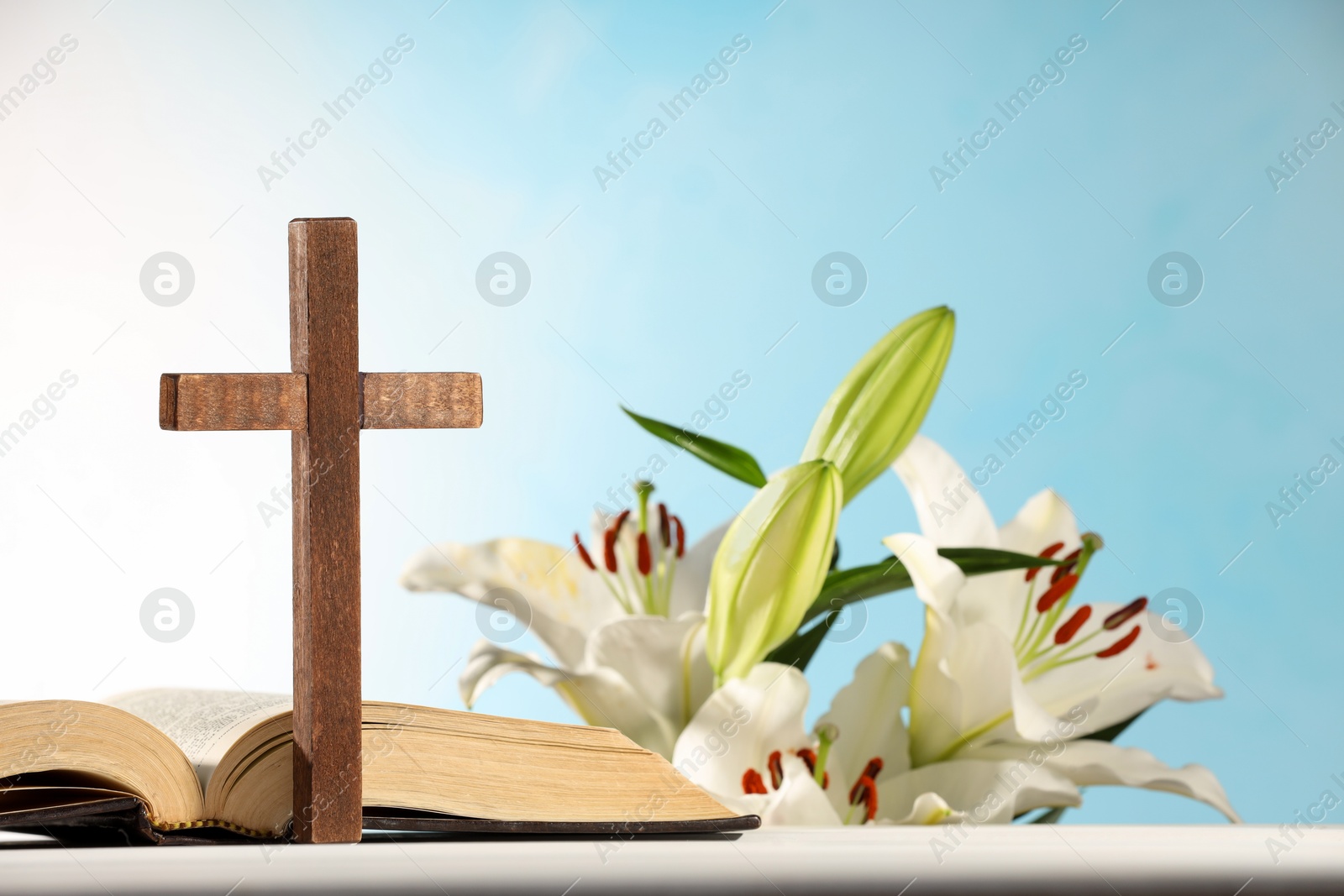 Photo of Wooden cross, Bible and beautiful lily flowers on white table against light blue background, closeup. Religion of Christianity