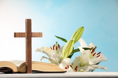 Wooden cross, Bible and beautiful lily flowers on white table against light blue background, closeup. Religion of Christianity