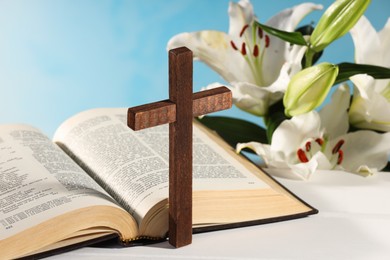 Photo of Wooden cross, Bible and beautiful lily flowers on white table against light blue background, closeup. Religion of Christianity