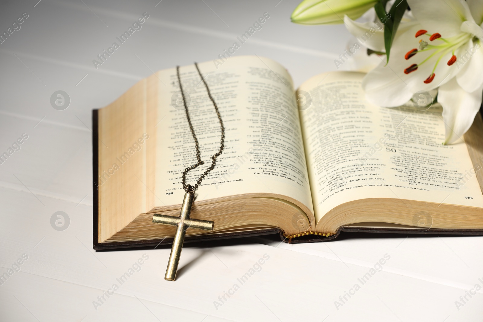 Photo of Cross with chain, lily flower and Bible on white wooden table. Religion of Christianity