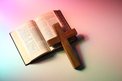 Photo of Bible and wooden cross on textured table in color lights, top view with space for text. Religion of Christianity